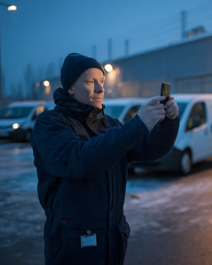 En mann i arbeidsklær tar bilde med telefonen på et depot i morgenmørket.