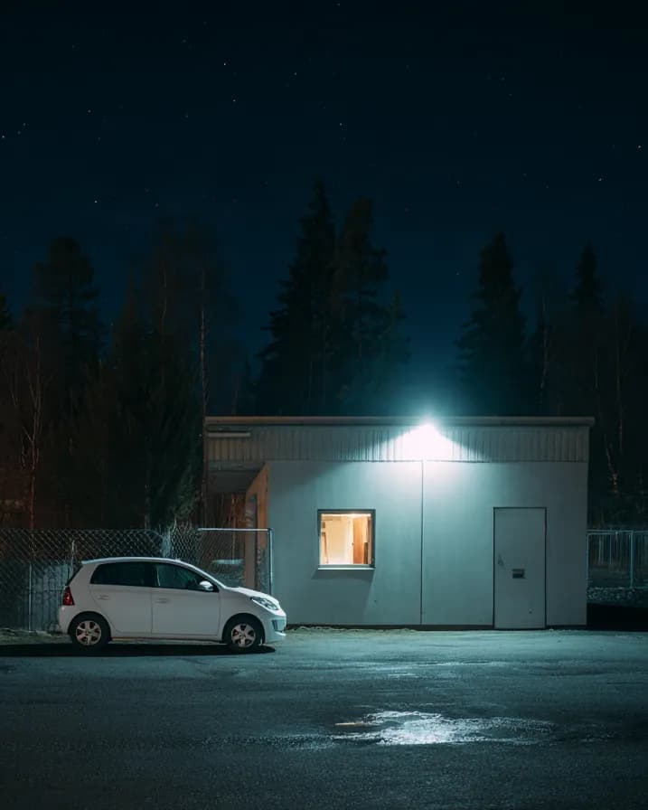 En hvit bil står alene ved et opplyst vaktbygg om natten.